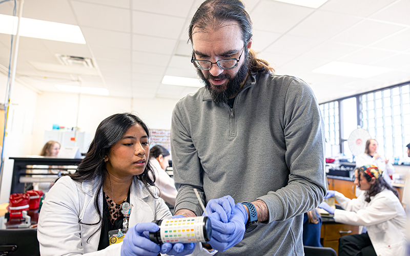A professor wearing a gray shirt and blue gloves holds a laboratory instruments.