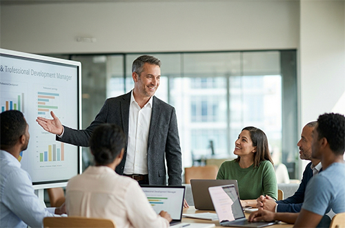 A manager standing near a digital display or whiteboard, mid-conversation while engaging with a small, attentive group of diverse adult professionals.